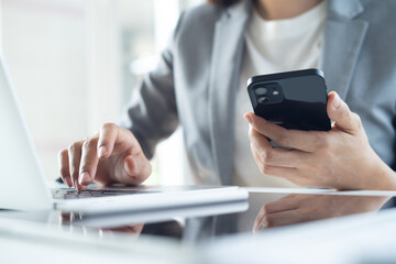 Close up, business woman using smartphone and working on laptop computer on office table. Businesswoman surfing the internet, web browsing via laptop and searching the information
