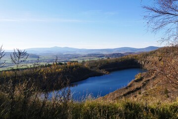 Panoramic view of serene blue lake surrounded by autumnal trees on rolling hills, vibrant landscape capturing stunning natural beauty under a clear sky with distant mountain range