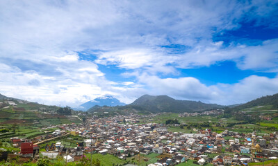 A scenic view of a town surrounded by lush green hills and mountains under a blue sky with clouds.