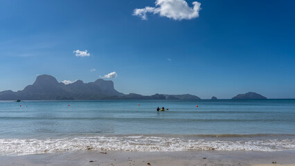 Beautiful seascape. A sunny day. The waves of the turquoise ocean are foaming on the sandy beach. Silhouette of a canoe and people in the water. A row of buoys in the distance. Picturesque mountains  
