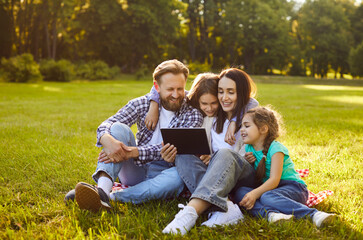 Happy family of four sitting on grass in park and watching something on tablet together. Smiling parents and children hugging while having fun and spending quality time outdoors on sunny summer day.