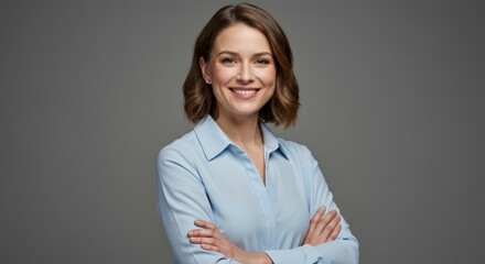 Confident young blonde woman with blue eyes smiling at the camera, standing with arms crossed against a gray background. She is wearing a light blue professional shirt.