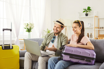 Happy young couple sitting on sofa with packed suitcases nearby, looking at laptop screen. Cheerful travelers in summer clothes planning vacation online, booking tickets and selecting tour together.