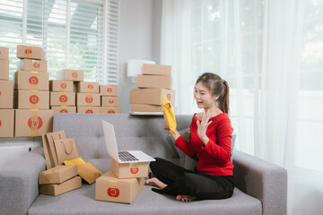 A young woman is comfortably working on a laptop on a sofa, surrounded by numerous shipping boxes,...