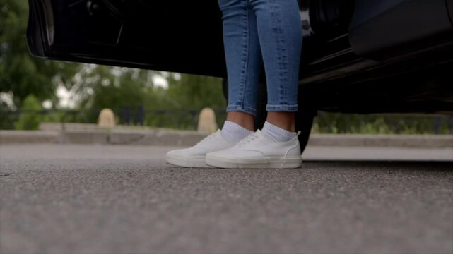 Close-up of female legs in trendy sneakers exiting vehicle and walking on parking lot in city street during road trip.
