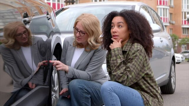 Upset black woman driver and female auto insurance agent assessing vehicle damage, discussing deductible, claims, coverage options and collision insurance after road accident in city.