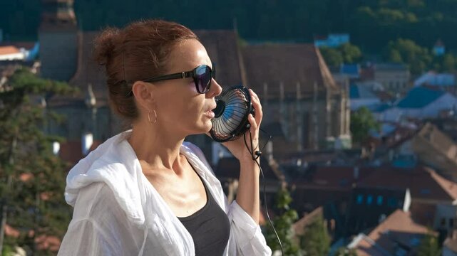 ReTourist using portable fan during heatwave. Redhead tourist wearing sunglasses and white shirt uses portable fan to refresh herself during heatwave with a cityscape in the background
