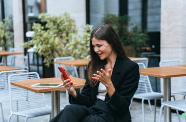Woman in black suit, excitedly interacting with a red smartphone, sitting at a chic outdoor café.