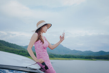 A young woman with closed eyes enjoys the breeze, leaning out of a car window against a scenic...
