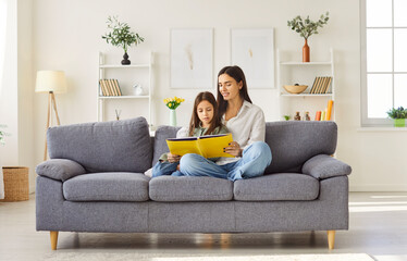 Young happy mother reading a book with her teenage daughter sitting on sofa at home. Smiling female parent spending time with child resting on couch on weekend. Leisure time together concept.