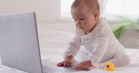 Portrait of cute little toddler child in white clothes sitting on bed in bedroom looking at laptop monitor screen playing with computer at home. Technology and family leisure concept. - Powered by Adobe