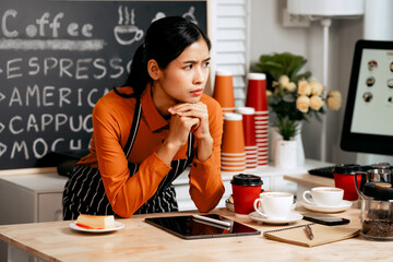 A young Asian female barista in a striped apron and orange shirt stands in a cozy modern café,...