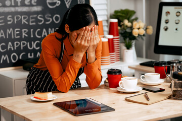A young Asian female barista in a striped apron and orange shirt stands in a cozy modern café,...