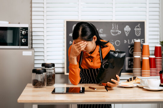 A young Asian female barista in a striped apron and orange shirt stands in a cozy modern café, looking sad, stressed, and disappointed, feeling exhausted and overwhelmed by business loss and failure.