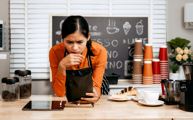 A young Asian female barista in a striped apron and orange shirt stands in a cozy modern café,...