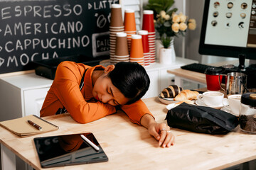 A young Asian female barista in a striped apron and orange shirt stands in a cozy modern café,...