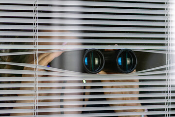 A man with binoculars observing through the blinds. The concept of surveillance, spying or covert surveillance.