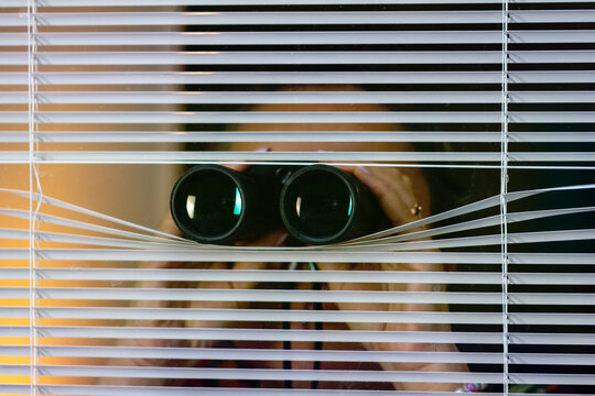 Woman peeking through blinds while holding binoculars. Ideal for illustrating themes of private detective or neighbourhood watch.