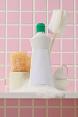 A large bottle of cleaning liquid stands beside white towels and yellow sponges, captured artistically in front of a warm pink tiled wall.