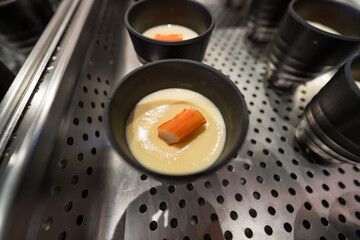 Close-up of a black cup filled with creamy custard topped with a piece of imitation crabstick, placed on a metal perforated tray