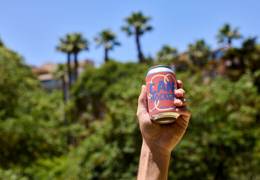 A hand raises a can joyfully, surrounded by lush greenery and bright palm trees under a sunny blue sky. PSD template mockup.