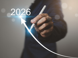 Person in dark suit holding white stylus pen drawing upward arrow pointing to year 2026,...