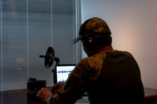 A man wearing camouflage clothing and headphones works at a laptop while listening to an audio file.