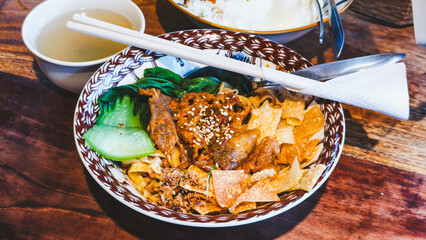 A vibrant bowl of spicy dry noodles with beef, greens, and crispy crackers paired with a saucy rice bowl topped with a fried egg—served with soup and a refreshing drink.