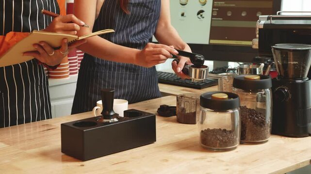 Two young Asian female baristas in striped aprons and orange shirts enjoy teaching each other how to make latte coffee, smell aroma, and taste coffee and croissants together in cozy modern caf&eacute;.