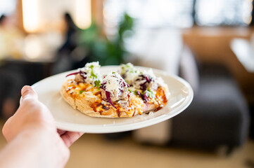 Close-up of a hand holding a plate with a freshly baked slice of pizza topped with melted cheese, onions, and herbs, against a blurred indoor background.