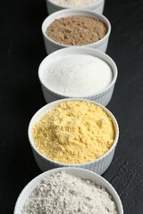 Different types of flour in bowls on black textured table, closeup