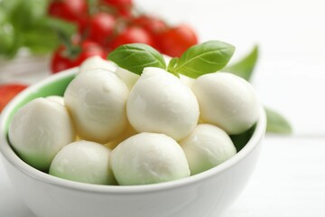 Tasty mozzarella cheese balls in bowl, basil and tomatoes on white table, closeup