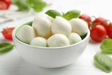 Tasty mozzarella cheese balls in bowl, basil and tomatoes on white wooden table, closeup