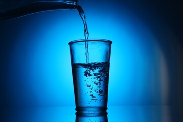 Pouring water from bottle into glass on blue gradient background. Backlit
