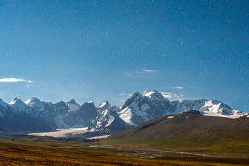 A wide shot capturing the Milky Way arching over a majestic range of snow-capped mountains and a vast, wild landscape at night
