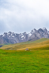 A small, rustic house sits isolated in a sprawling green mountain meadow, with snow-capped, jagged mountains forming a dramatic background