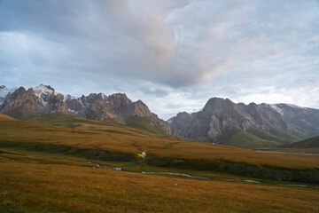 A broad view of a mountain landscape at twilight, with faint vehicle lights, a winding river, and distant snow-capped peaks under a cloudy sky
