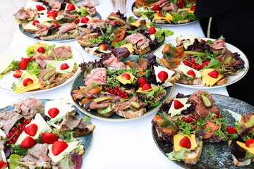 Colorful open-faced sandwiches with assorted meats, cheeses, lettuce, strawberries, and red currants arranged on rustic bread, served on decorative plates.