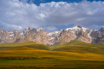 Broad golden and green pastures stretch towards a majestic range of snow-capped mountains, under a...