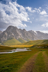 A small river winds through a sweeping green and golden mountain valley, leading towards towering mountains under a cloudy, bright sky