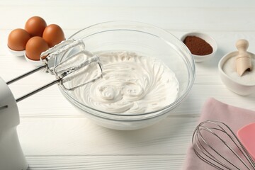 Bowl with whipped cream, hand mixer, whisk and ingredients on white wooden table, closeup