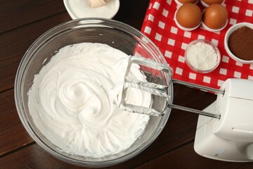 Bowl with whipped cream, hand mixer and ingredients on wooden table, flat lay