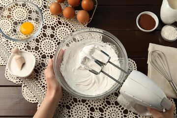 Woman whisking cream with hand mixer at wooden table, top view