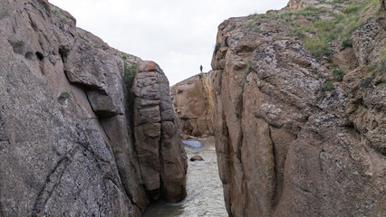 A lone adult person of unknown race stands precariously on a cliff edge, looking down into a narrow canyon with a river flowing between rock walls