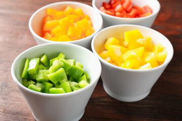 Pieces of fresh colorful bell peppers in bowls on wooden table, closeup