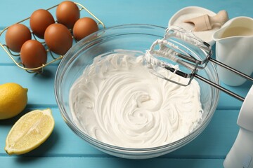 Hand mixer with whipped cream, eggs and other products on light blue wooden table, closeup