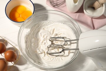 Hand mixer with whipped cream, eggs and other products on white wooden table, flat lay