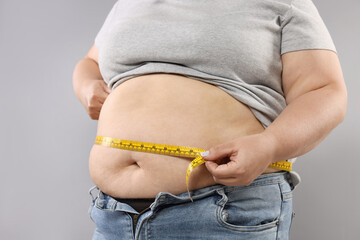Overweight woman measuring her belly with tape on grey background, closeup