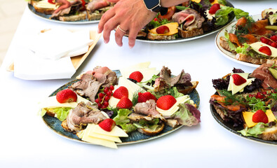 Colorful plate of open-faced sandwiches with cheese, cold cuts, lettuce, strawberries, and red currants served on a white tablecloth at a catering event.