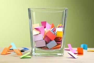 Jar with colorful pieces of paper on wooden table against green background, closeup